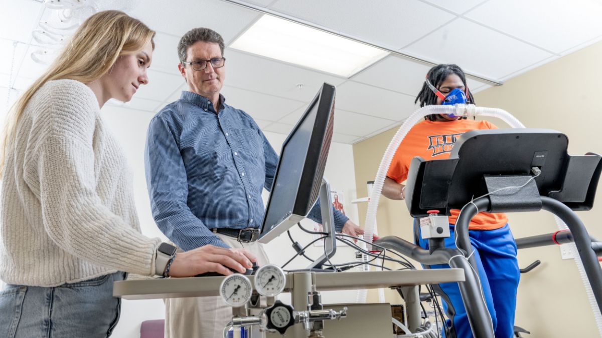 A student operating a fitness testing device while another person exercises on a treadmill with respiratory monitoring equipment.