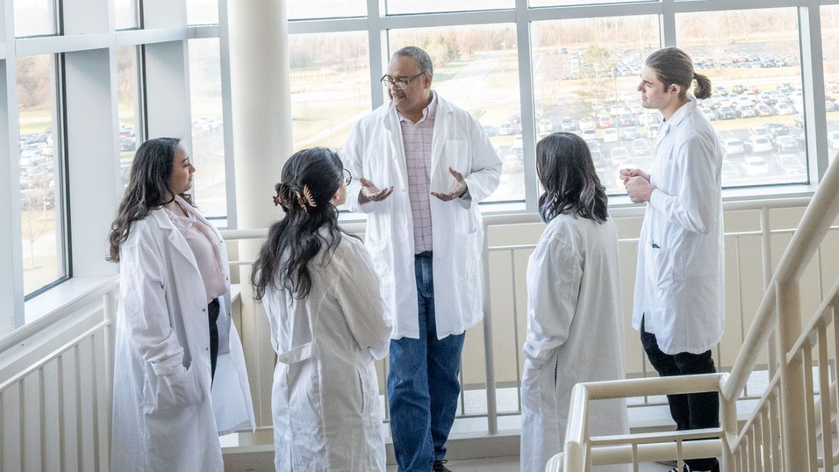 A professor mentoring students in lab coats in a bright staircase area.