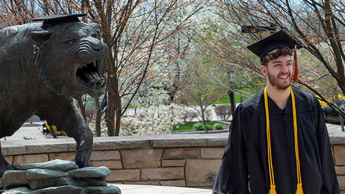 a student wearing a cap and gown stands next to the Tiger statue in front of a brick wall