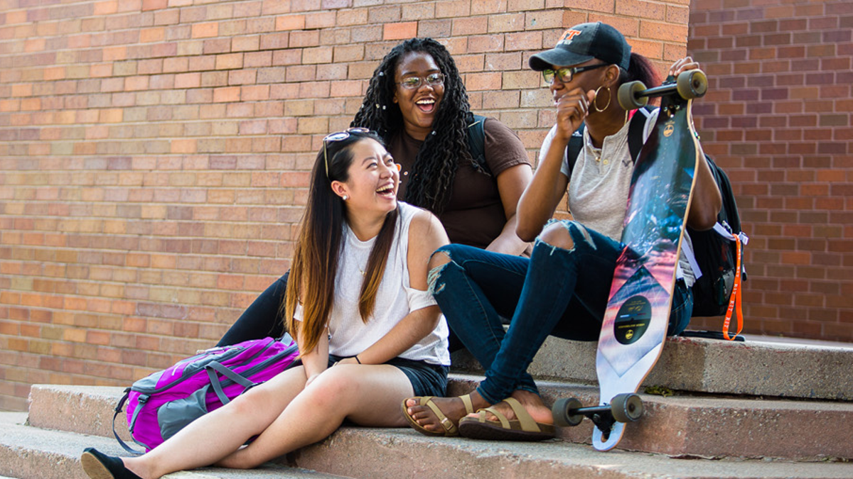 Three students sit on steps, one holding a skateboard, enjoying a casual moment together outdoors.