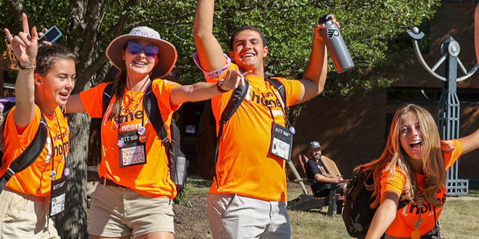 ﻿Four young adults in orange welcome home tiger shirts and backpacks, gathered together, exuding excitement  with hands raised and smiling