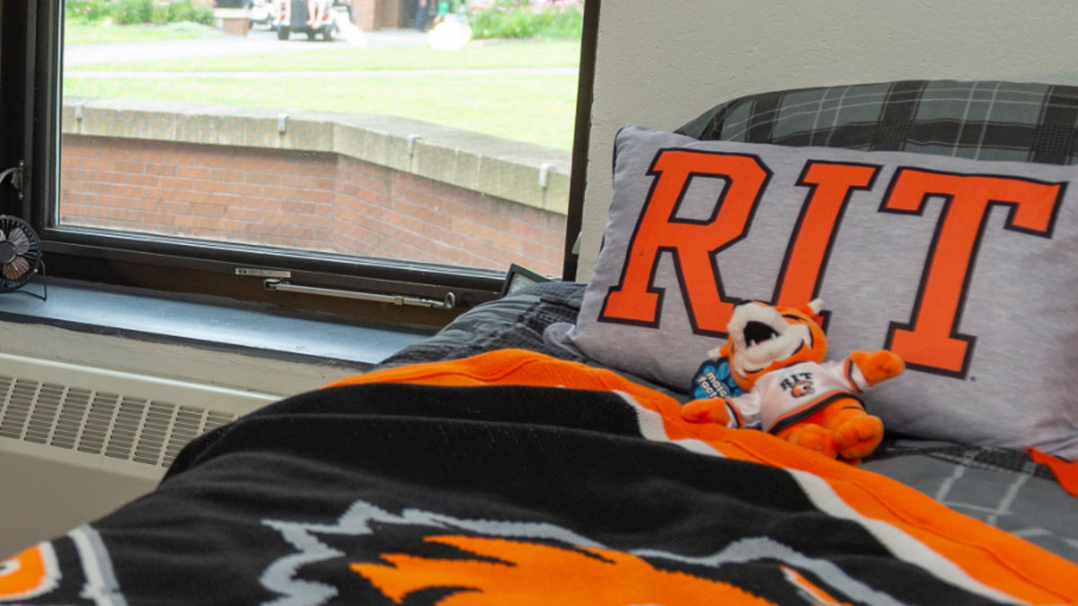 A neatly made bed featuring a pillow and blanket, prominently displaying the word "RIT" on the pillow.