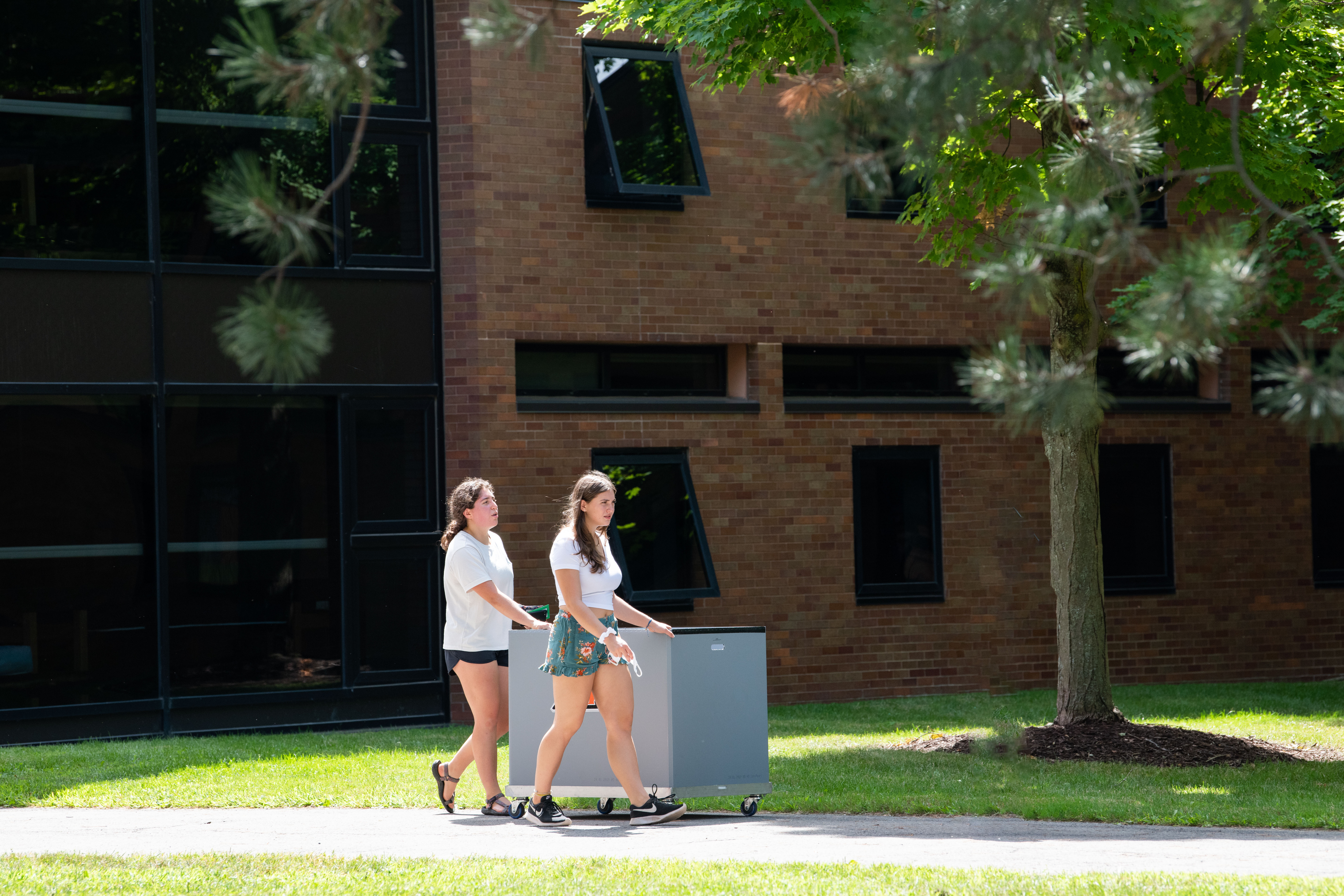 2 female presenting students push a housing cart outside in the sun