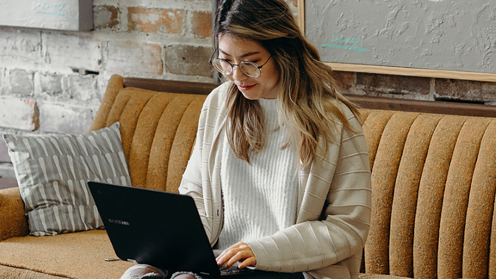 woman in white sweater sitting on a yellow sofa, typing at a laptop