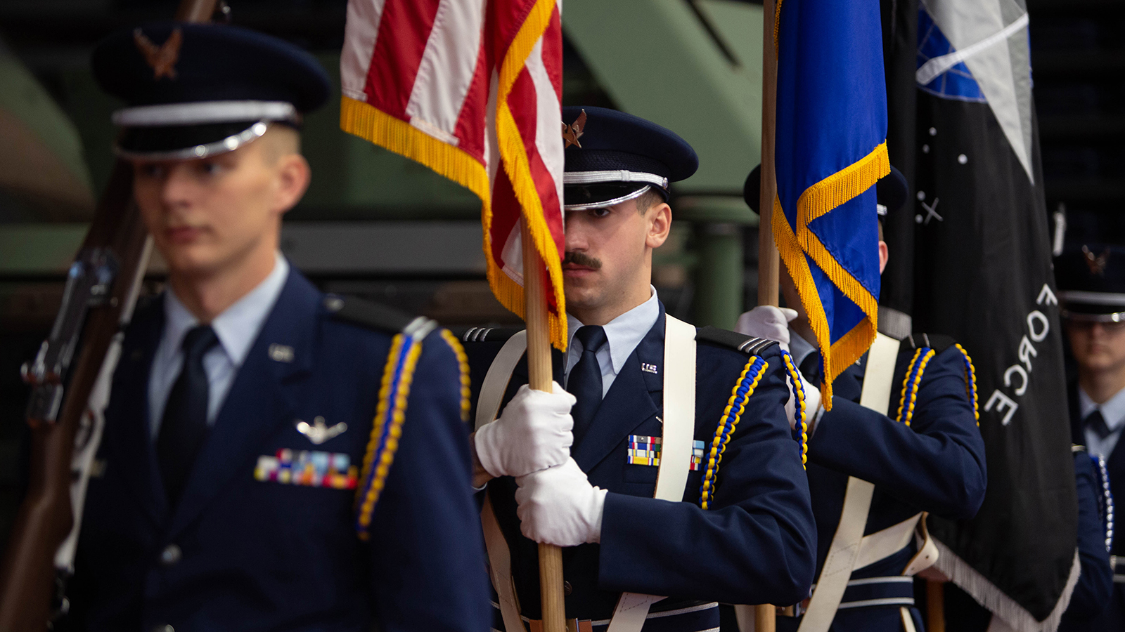 A group of individuals in military uniforms participating in a flag ceremony. Three flags are being held upright, including the United States flag.