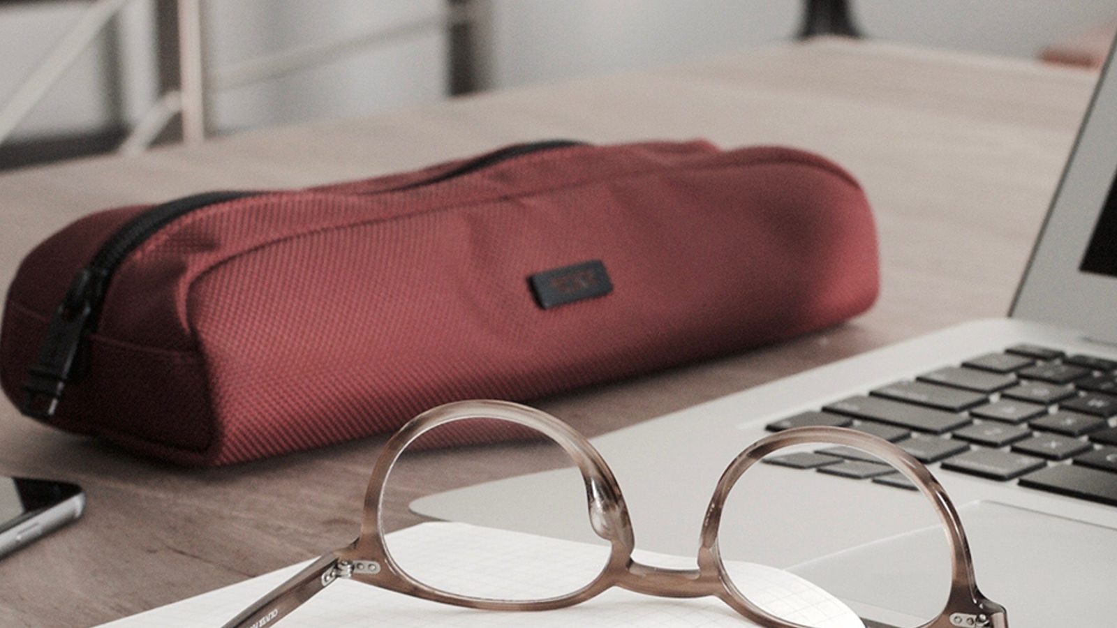 angled photo of a laptop, glasses, phone, and a red pencil holder on a desk