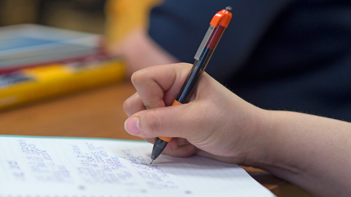 A hand holding pen writes on lined paper on desk.