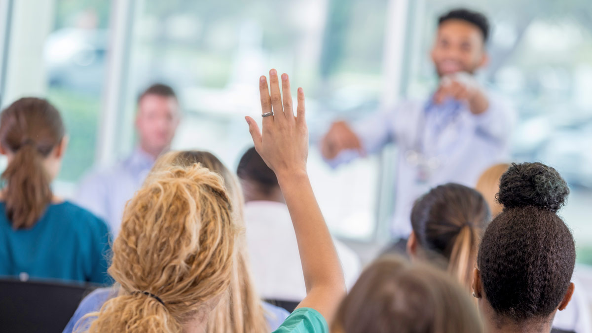 Close up of a hand raised in a classroom with a teacher out of focus in the background..