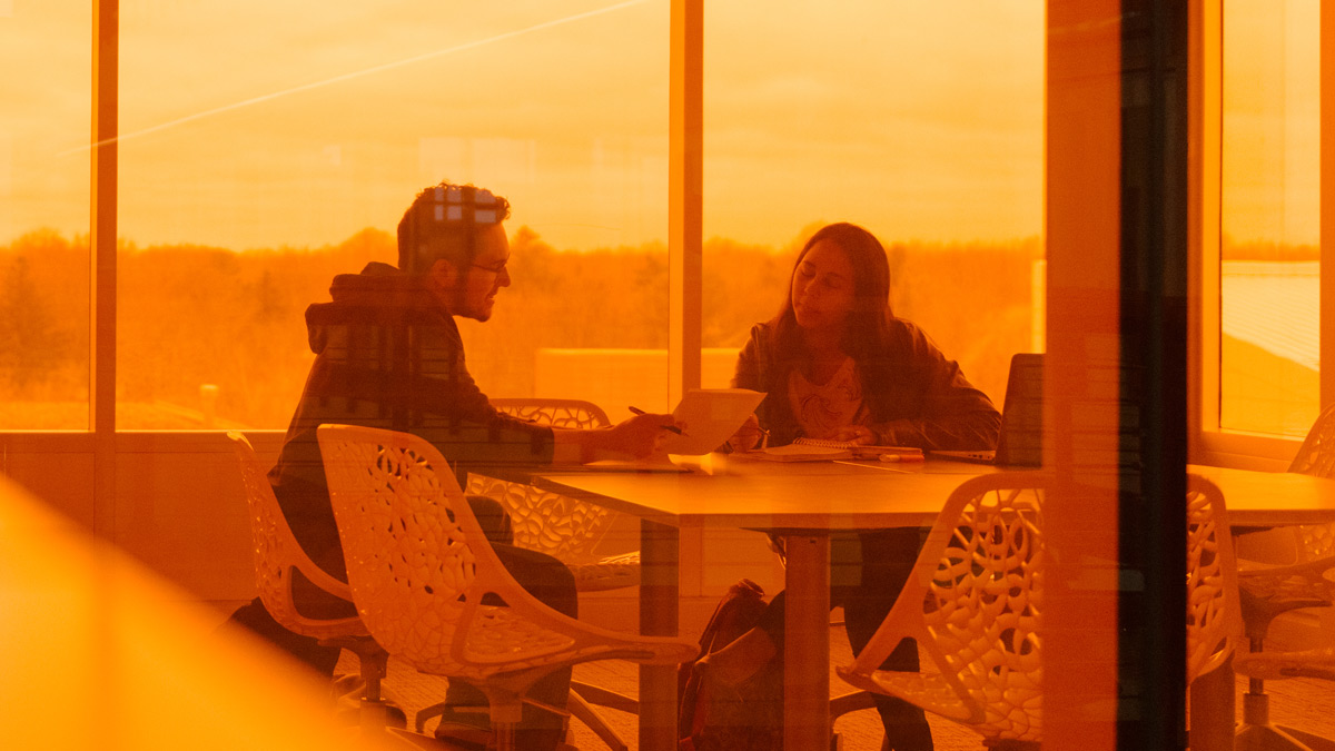 Photo taken through orange glass of two students sit in a conference room looking over a paper.