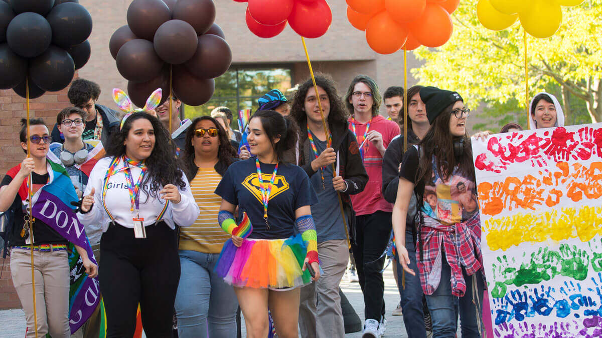 Students marching with rainbow-colored balloons and a white banner with handprints of different colors.