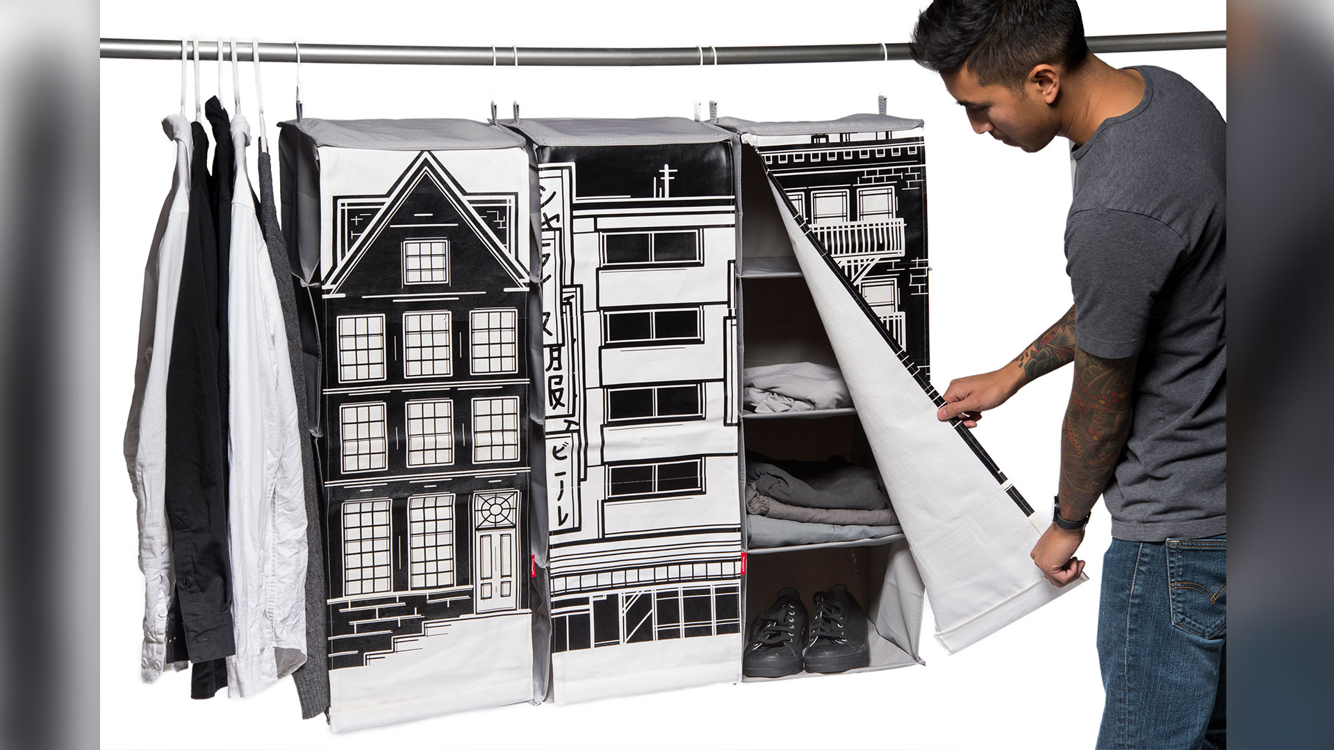 Front view of a series of 3 closet organizers hanging on long pole, each designed to look like city building facades in black and white, student opening front flap of organizer to show 3 interior vertical shelves with assorted clothing items folded
