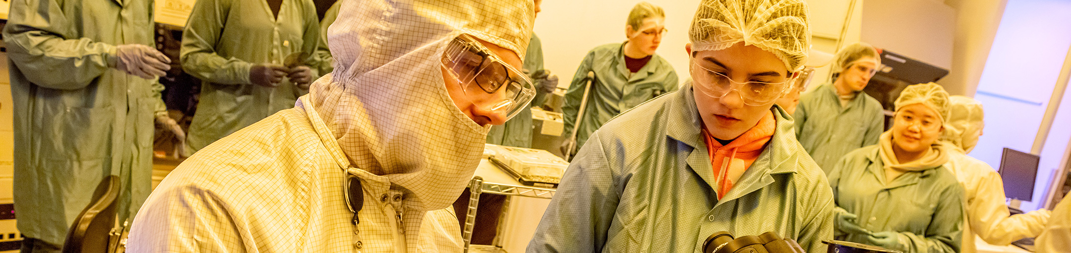 Students and faculty in a cleanroom, looking at a microscope.