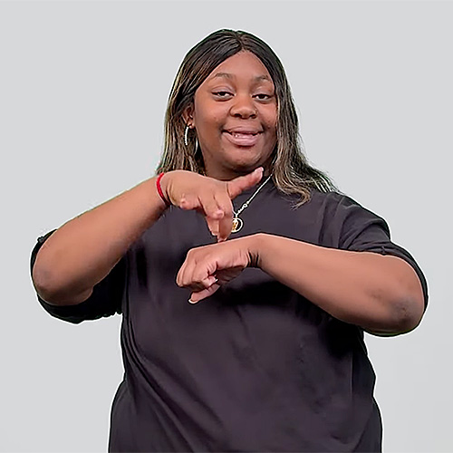 The image shows a person with long, dark brown hair that has light brown highlights. They are wearing a loose-fitting black shirt and have a red bracelet on their right wrist. The person is smiling and making a hand gesture with their right hand pointing downward and their left hand positioned horizontally, palm facing down. They are signing 'pumpkin.' The background is plain and light gray.