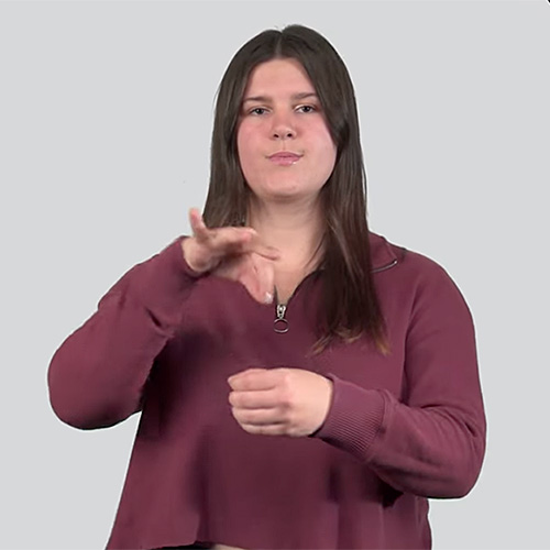 A person is shown with long, straight brown hair. They are wearing a maroon colored long sleeve shirt. The person's lips are pursed and is making a hand gesture with their left and right hands, making the sign for 'ghost.' The background is plain and light grey.