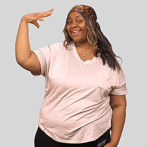 The image shows a smiling woman standing against a light gray background. She has long, wavy, dark hair with light streaks and wears a leopard-print headband. She is wearing a light-colored, short-sleeve T-shirt and black pants. Her left arm is raised, with her palm facing towards and fingers slightly curved, creating the sign for ‘sun.’ 