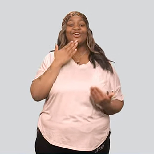 The image shows a smiling woman standing against a light gray background. She has long, wavy, dark hair with light streaks. She is wearing a light-colored, short-sleeve T-shirt and black pants. Her left arm is raised near her mouth, and her right hand is raised to her chest, creating the sign for 'grateful.' 