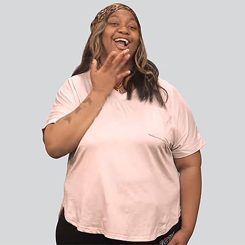 A woman is smiling with her left hand raised, fingers spread out, making the sign for ‘colors.’ She has long hair with light-colored streaks and is wearing a light pink T-shirt and black pants. She is standing against a plain light gray background.