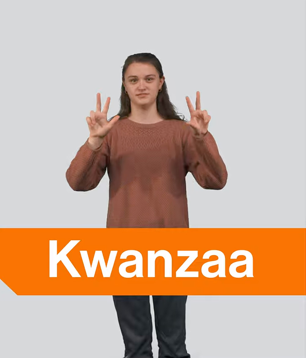 The image features a person standing upright, wearing a long-sleeved, reddish-brown sweater and dark pants. They are holding up both hands with their thumbs, index, and middle fingers extended to form the sign for ‘Kwanzaa’. The person has a neutral facial expression with a hint of a smile and long, dark hair hanging loose. Across the bottom of the image, there is an orange banner with bold white text that says "Kwanzaa." The background is a plain light gray.