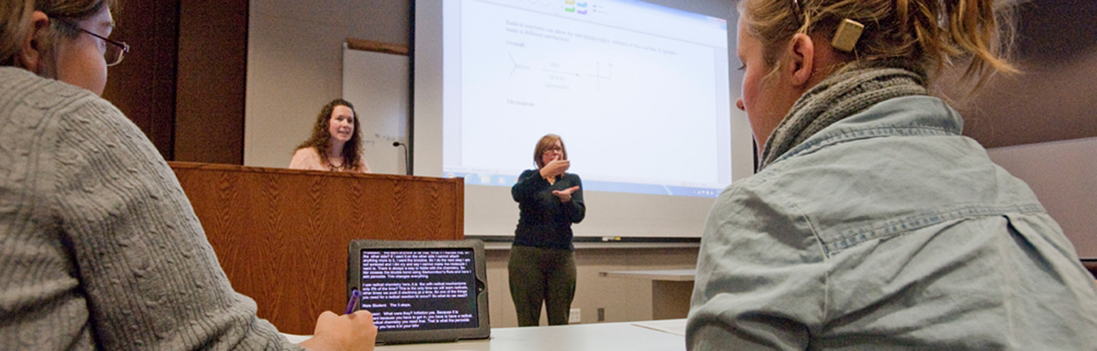 Two people seated at a table with captions displaying on an iPad, while an interpreter signs for the lecturer at the front of a classroom.