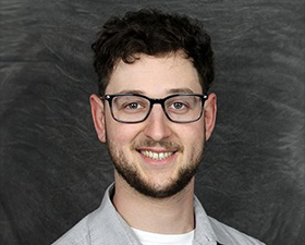 Young man, dark hair, dark glasses grey open collar shirt over white t-shirt.