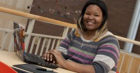 Young woman, dark skin, wearing striped sweater, sitting at table with laptop, stairway in background.