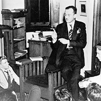 Early b/w photo of Ed Scouten in room with bookcase and books stacked around, sitting on arm of chair and speaking to students seated on floor and looking up at him