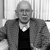 B/W photo of Ed Scouten seated on couch in front of curtains, wearing plaid jacket with vest and bowtie