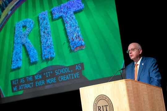 RIT President Munson stands at podium and addresses RIR community. A slide with students spelling out RIT is behind him.