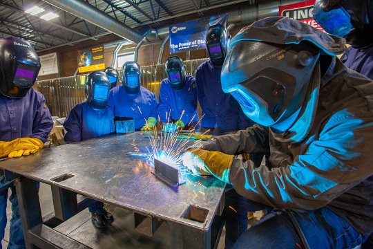 Eight people wearing welding gear stand around a table. One person is making sparks. 
