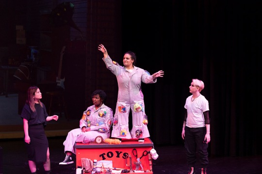 Four students acting on stage: one student is sitting on a red toy box and one student is standing on it. Two students stand on each side of the box.