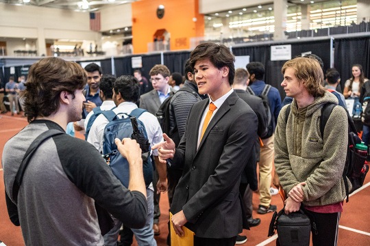 Male student who dressed in a black suit with a white shirt and orange tie is standing talking to another student who is wearing a black t-shirt. More students are in the background meeting with employers at the RIT Career Fair.