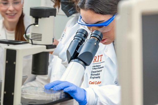 Female student wearing blue goggles, blue gloves, and white lab coat looks through a microscope.