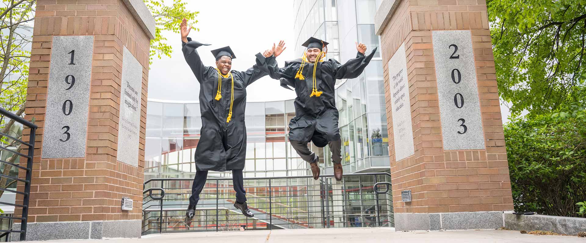 two recent grads wearing regalia jumping in the air in celebration.