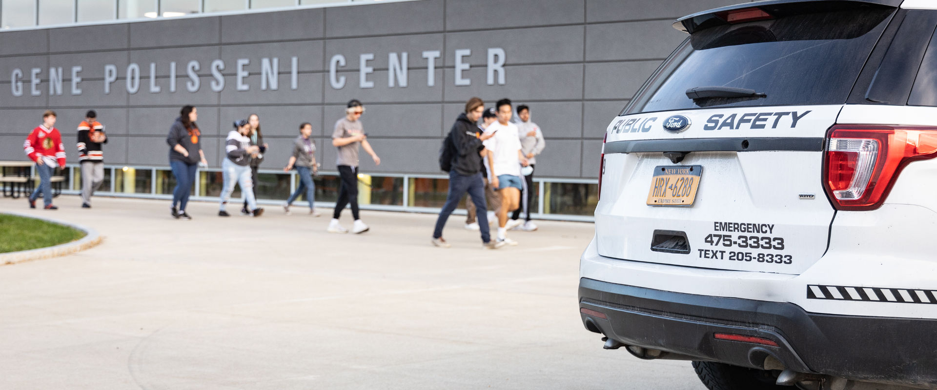 Group of individuals walking in front of the Gene Polisseni Center, with a parked emergency vehicle displaying contact information for emergencies.