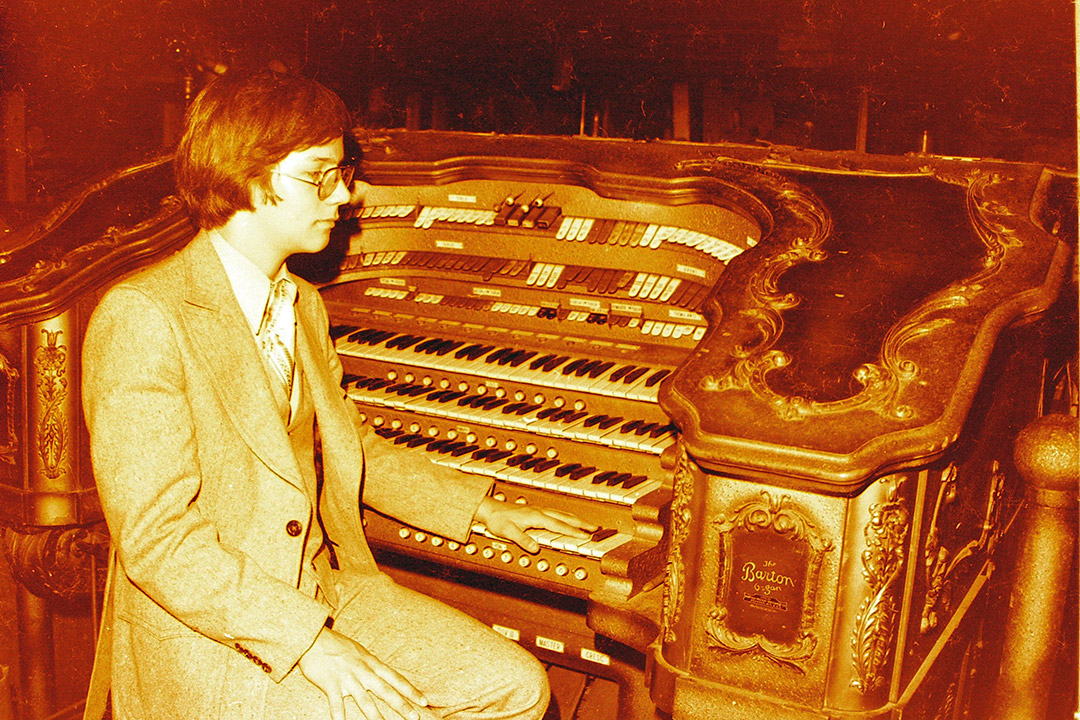 An old, amber colored photo shows a man in mid-century style clothing sitting in front of a pipe organ.