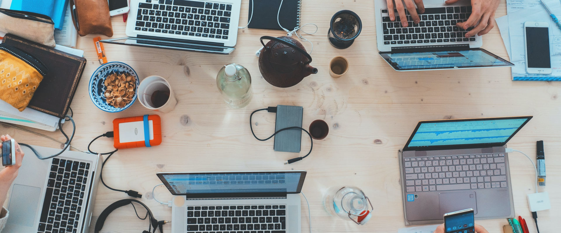 Overhead view of a busy work table with multiple laptops, notebooks, smartphones, and various personal items, indicating a collaborative working environment.