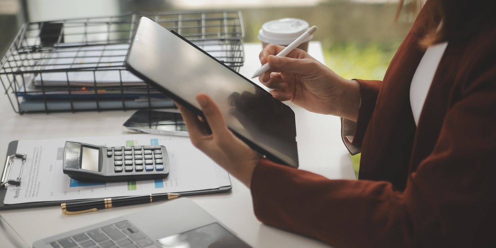 ﻿A female worker at her workstation holding a tablet and pen over her desk of  paperwork, a laptop, and a calculator