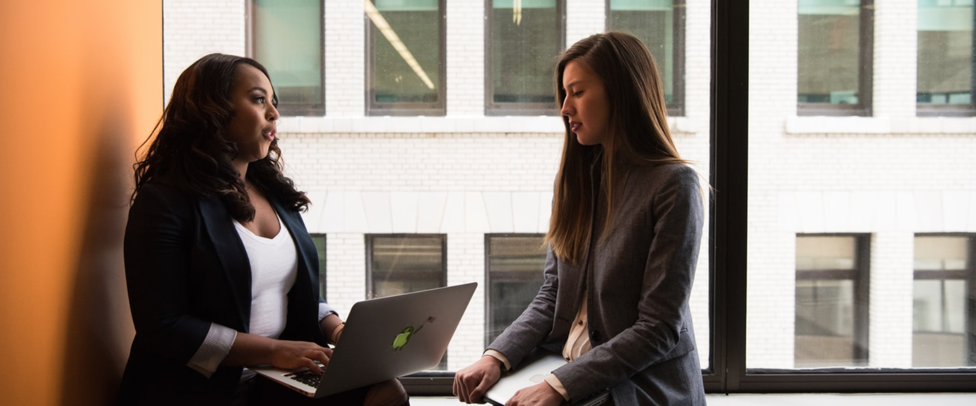 Two individuals with obscured faces sitting opposite each other at a table, one holding an open laptop with a green sticker on the cover, in a room with large windows overlooking buildings.