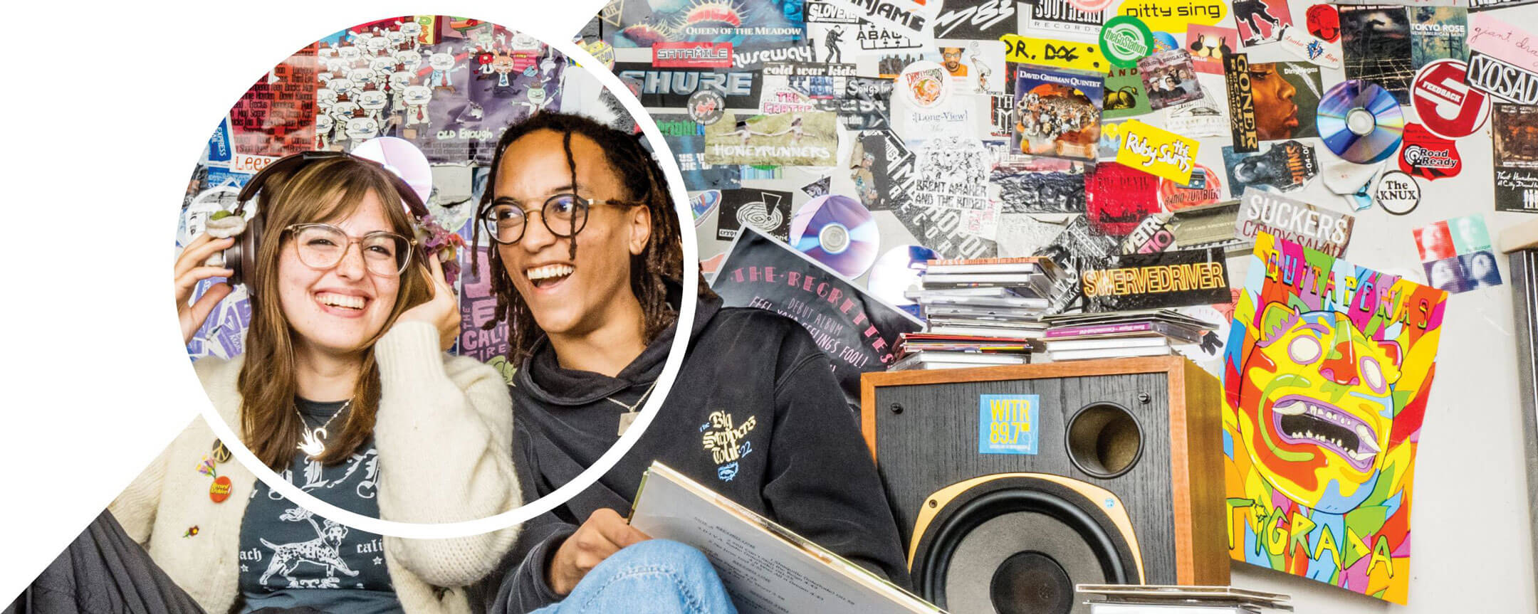 Julianna Upham and Sejjemba Magoye sitting together and smiling in front of a wall of stickers and a speaker, while listening to a vinyl record.