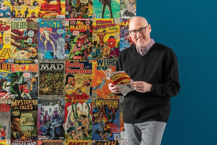 man holding a comic book leaning against a wall with a collage of varying comic book covers