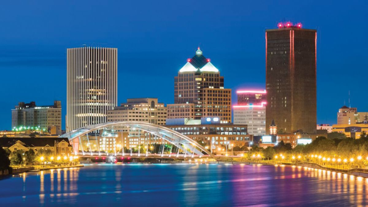 The skyline of downtown Rochester, New York, featuring illuminated skyscrapers and a bridge over a river at dusk.