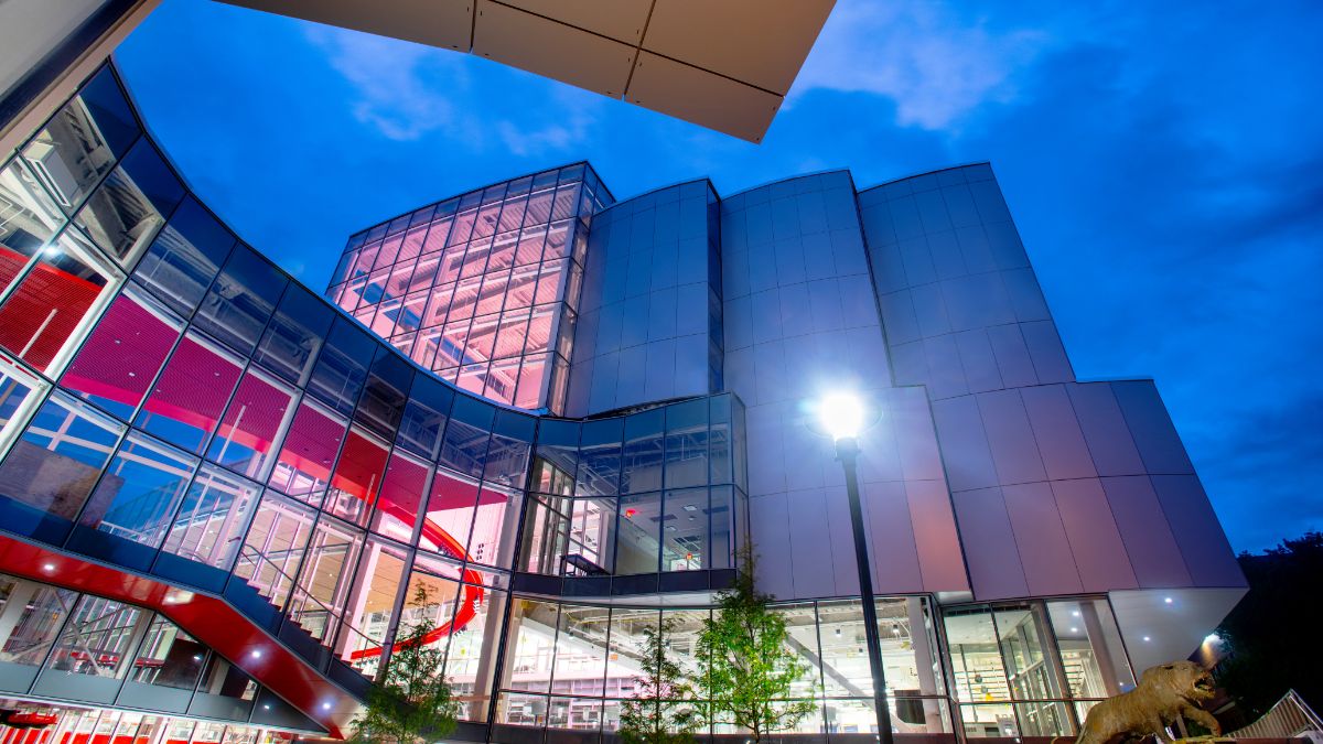 Glass-walled academic building with vibrant red staircases seen through the exterior at dusk.