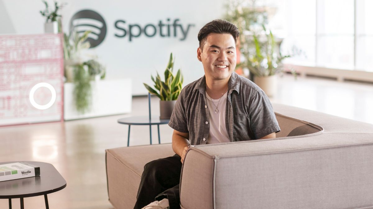 A young professional smiling while seated in an open, modern office space with a Spotify logo in the background.