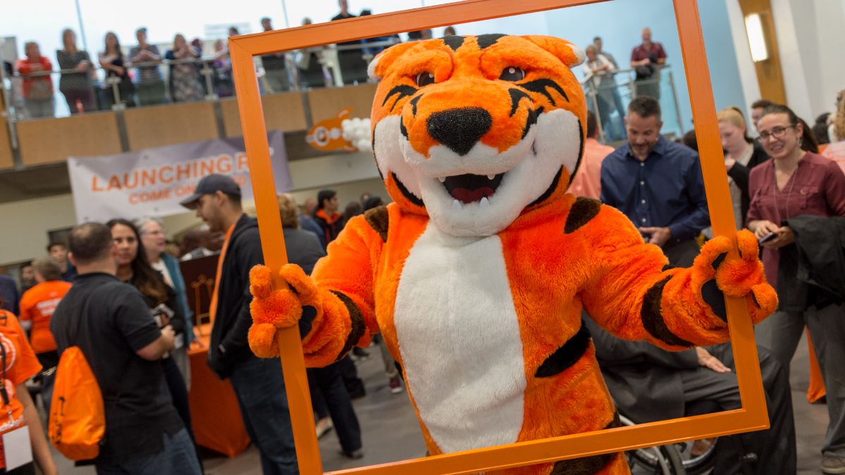 An orange tiger mascot posing with an orange frame at an event, surrounded by people and festive decorations.