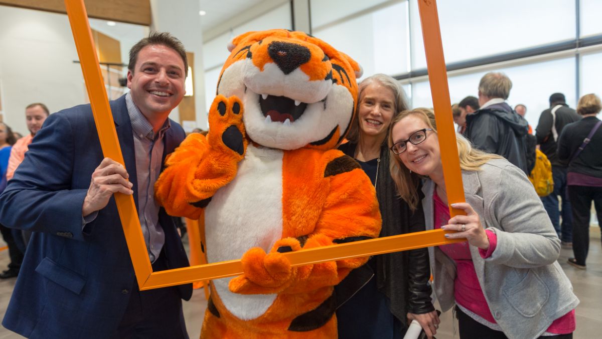 The orange tiger mascot posing with three smiling adults, holding an orange frame at a lively campus gathering.