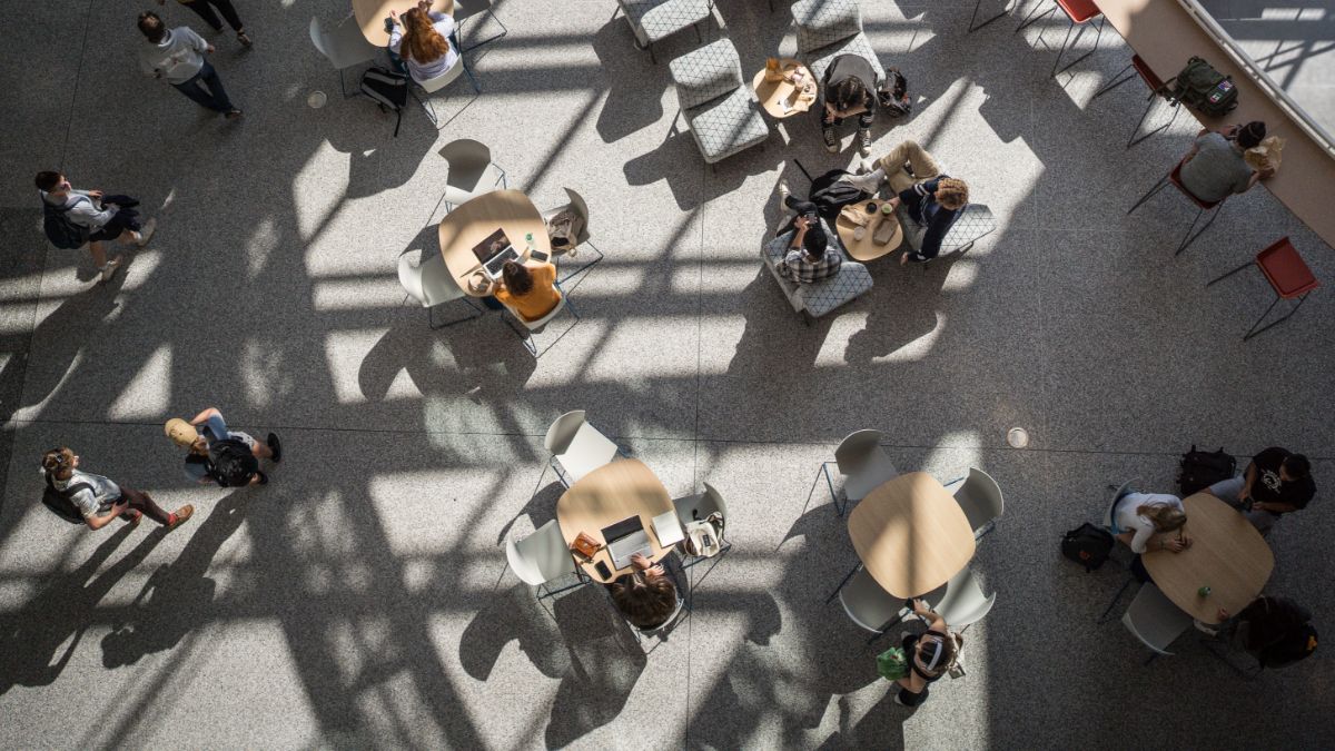 A bird's-eye view of students and staff sitting and conversing around tables in a bright, sunlit common area.