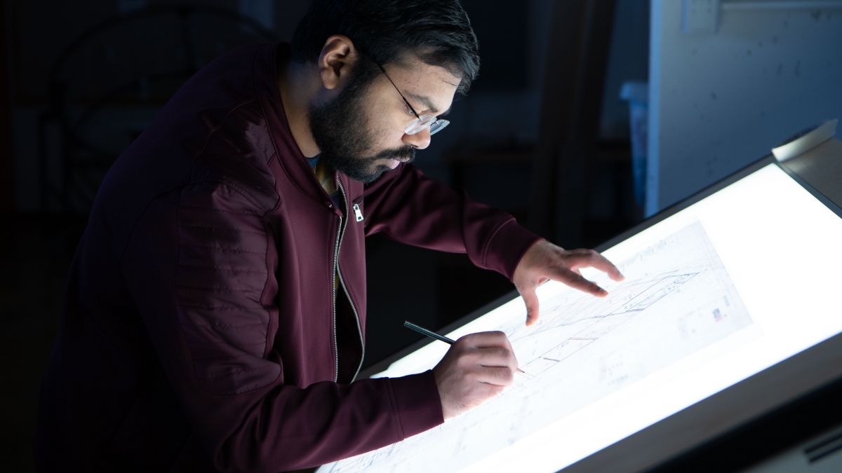 A student intensely working on architectural drawings illuminated by a light table.