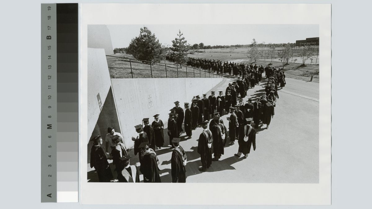 A black-and-white historical images showing two lines graduates lined up outside waiting to enter a field house.