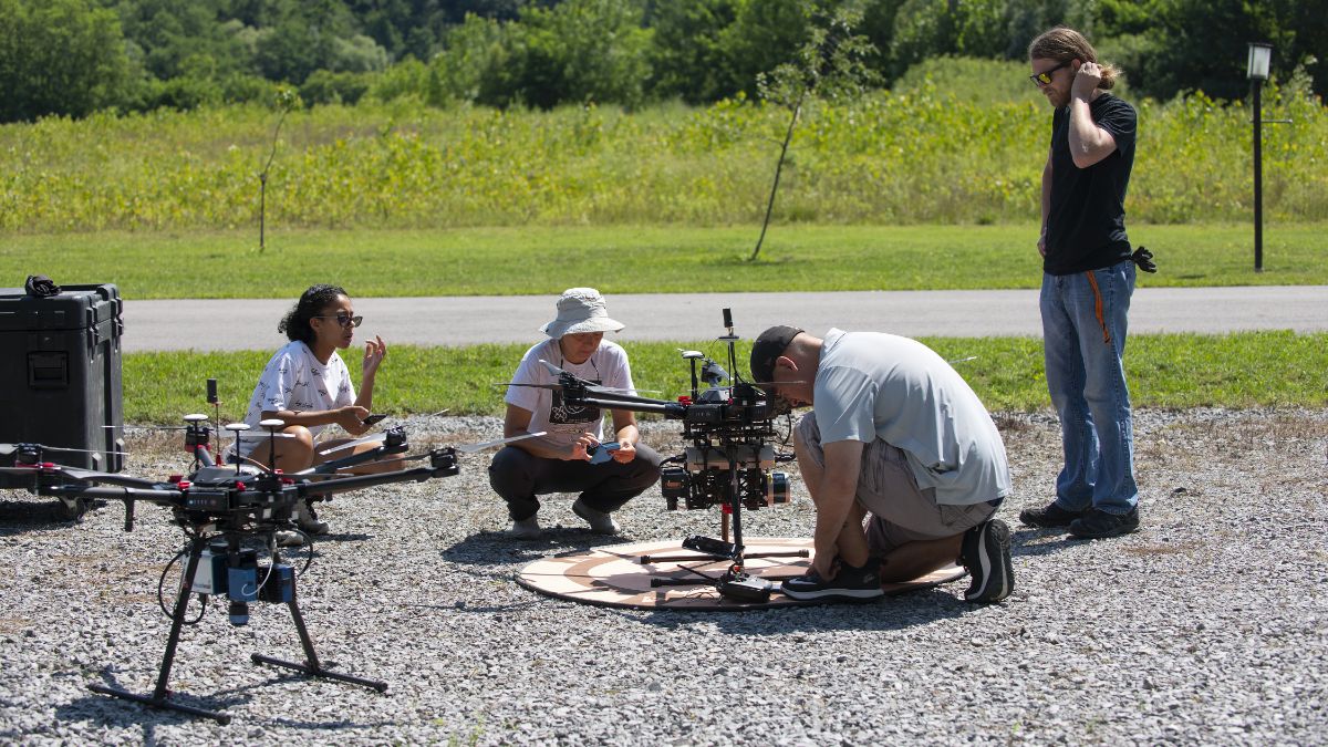 A group of people preparing drones for outdoor testing in a field on a sunny day.