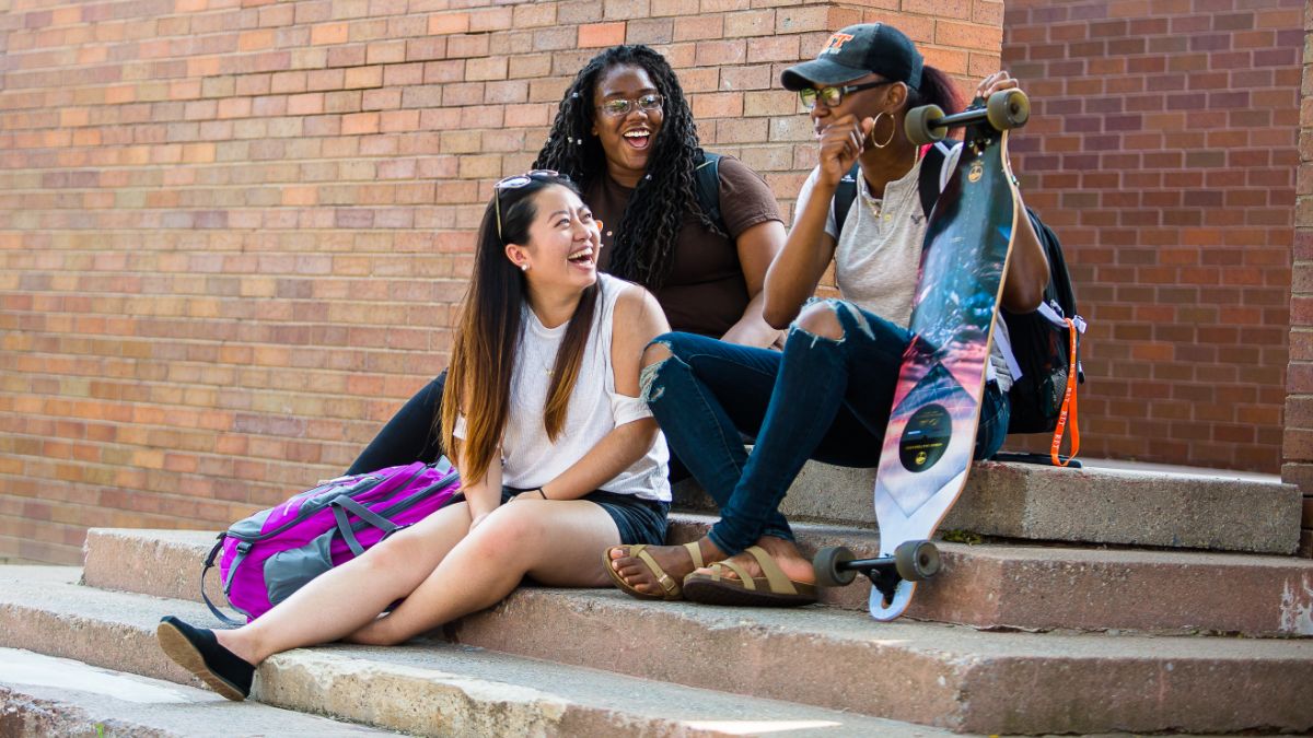 Three students sitting on steps outside, chatting and laughing together.